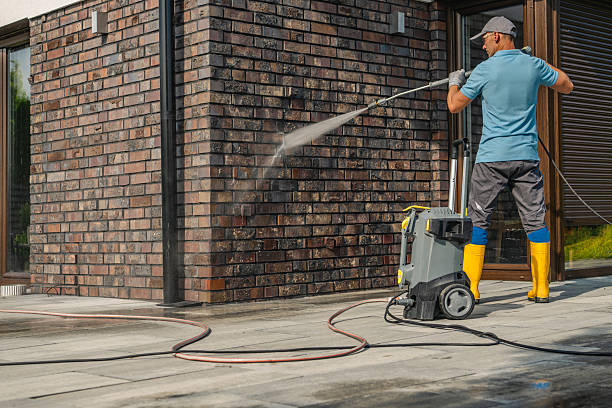 Man using an electric pressure washer to clean a dirty brick house wall.